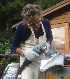 Josée de Vérité in front of a wooden cabin, polishing a piece of aluminum from a plane that crashed on Mont Blanc. She is wearing protective goggles.